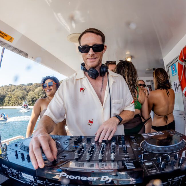 A DJ wearing sunglasses and a white shirt operates a Pioneer DJ controller on the deck of a private yacht charter in Sydney Harbour while several people dance and enjoy the sunny weather around him.