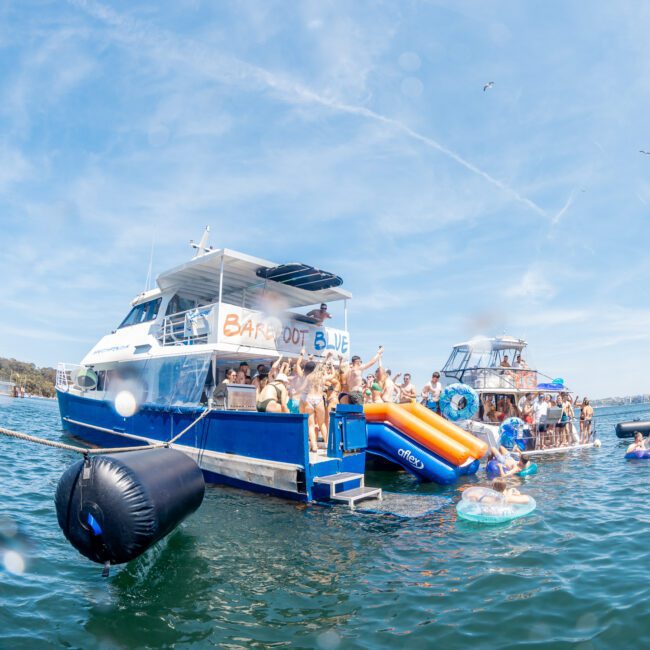 A group of people are enjoying a sunny day on "Barefoot Blue," a private yacht charter in Sydney Harbour. Some are on the deck, while others are in the water with inflatables. Several boats are nearby, creating a vibrant atmosphere ideal for corporate boat events in Sydney.