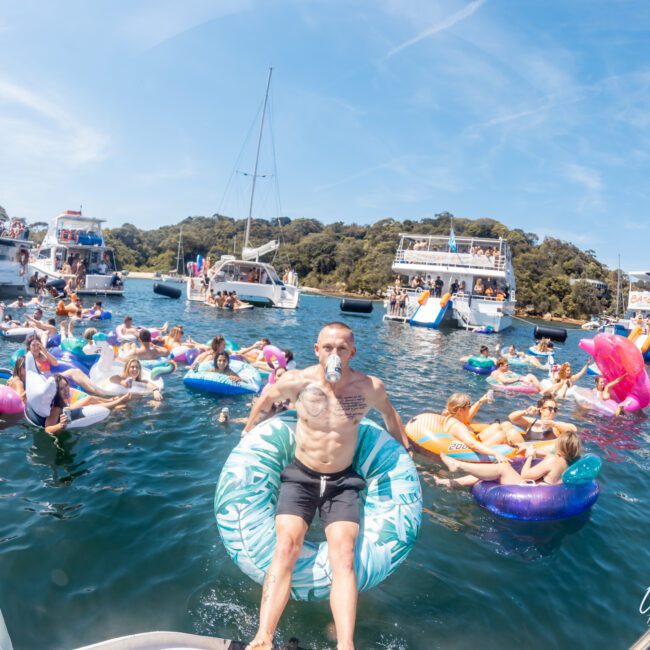 People on inflatable tubes and floats enjoy a lively boat party on a sunny day, surrounded by other boats and lush green trees in the background. Experience the fun with a luxury yacht hire Sydney for unforgettable moments.