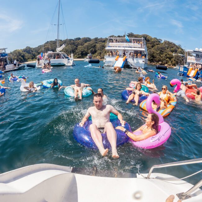 A group of people enjoys a sunny day on water inflatables near anchored boats in a scenic bay surrounded by green hills, making the most of their Sydney boat party hire.