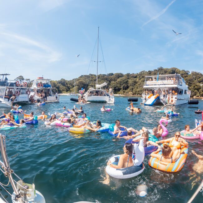 People relaxing on colorful inflatables in the water, surrounded by boats on a sunny day near a green, wooded coastline, enjoying a luxurious yacht hire in Sydney.