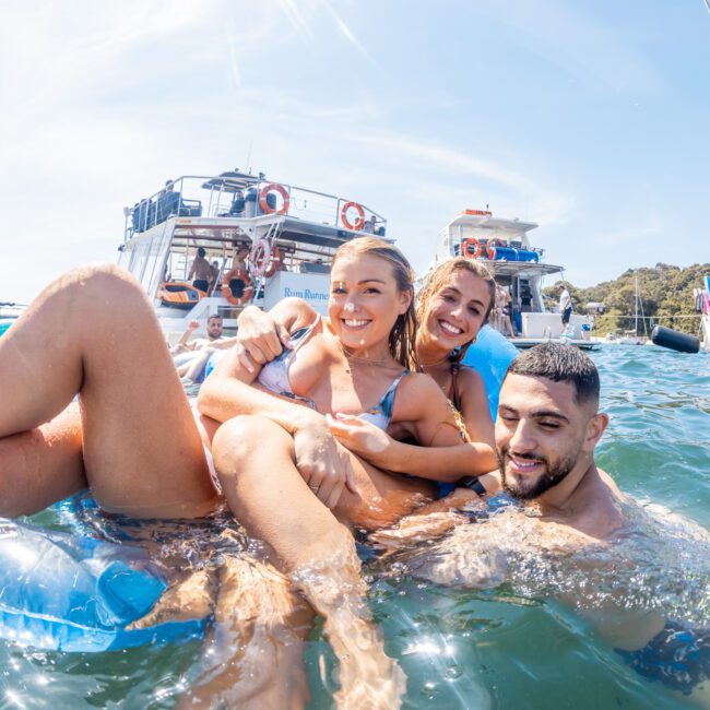 Three people in swimwear enjoy floating in the water near anchored boats on a sunny day, surrounded by others relaxing and swimming, making the most of their Sydney boat party hire.