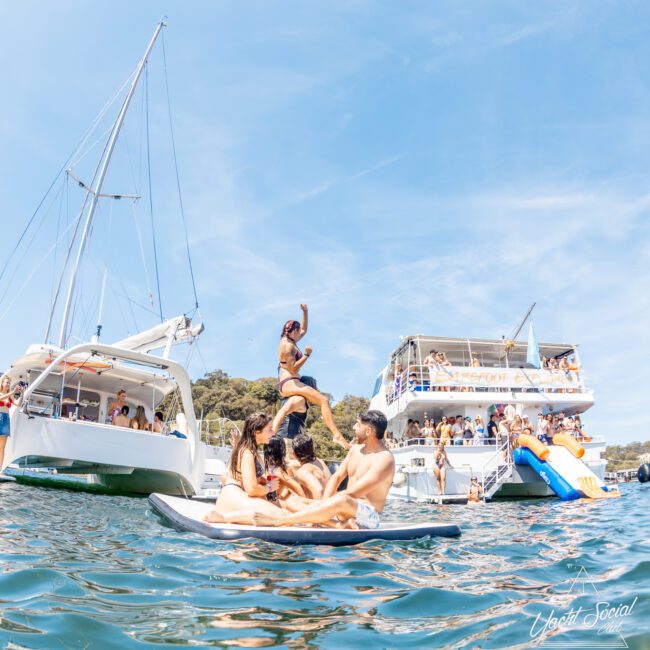 A group of people are enjoying water activities next to anchored boats. One person jumps into the water from a floating mat while others watch. A large boat with a slide is in the background as part of a corporate boat event in Sydney. The sky is clear.