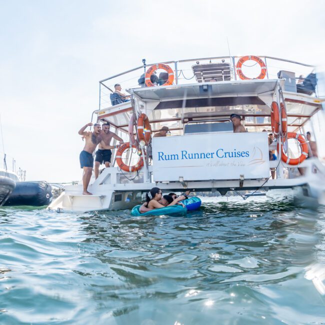 People swim and relax near a boat labeled "Rum Runner Cruises" on a sunny day, epitomizing the allure of a luxury yacht hire Sydney experience.