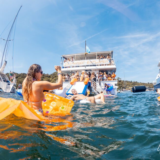 People are enjoying a sunny day on the water with private yacht charters on Sydney Harbour and inflatables, including a woman in an orange inflatable chair waving towards others on a boat.