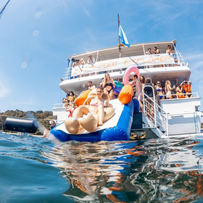 A group of people on a private yacht charter in Sydney Harbour are enjoying a sunny day, with several individuals seen on an inflatable float in the water and others on the boat's deck.