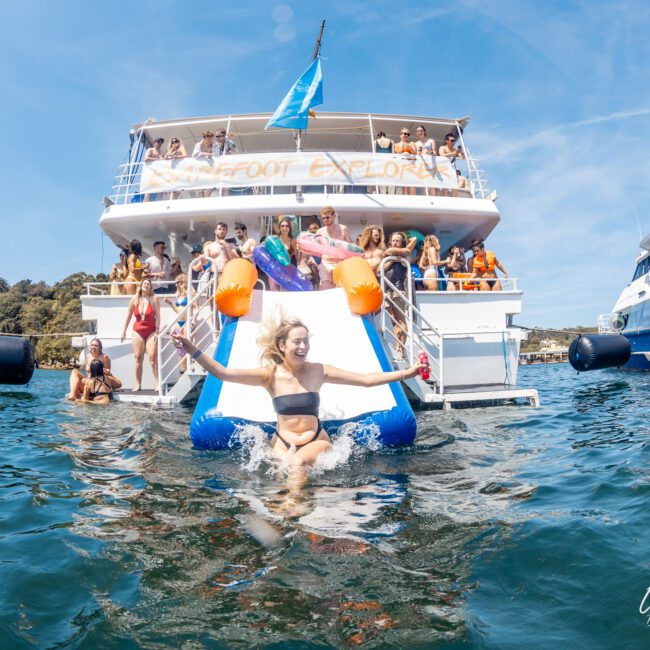 A woman slides down an inflatable slide into the water from a boat with "Barefoot Explorer" written on it, surrounded by people enjoying a private yacht charter in Sydney Harbour. Another similar boat is visible in the background.