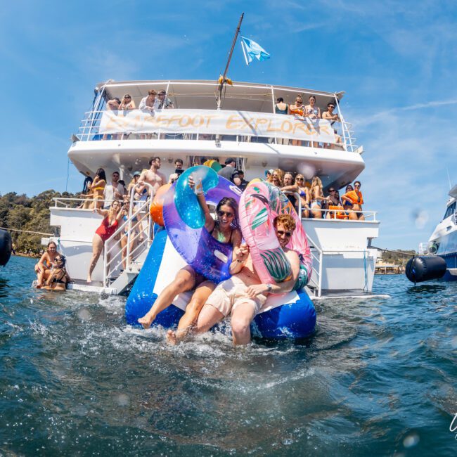 People slide into the water from the back of a yacht, surrounded by others on the boat. Inflatable floats and banners are visible, adding to the festive atmosphere. Another yacht is anchored nearby under the sunny Sydney sky, ideal for a Luxury yacht hire Sydney experience.