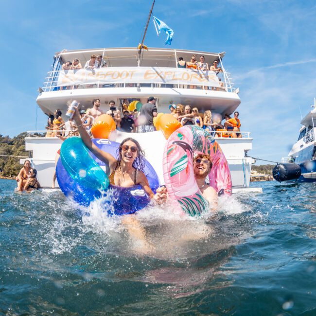 Two people in colorful swimsuits jump into the water from a boat during a corporate boat event in Sydney, with others on the luxury yacht watching and taking photos in the background under clear blue skies.