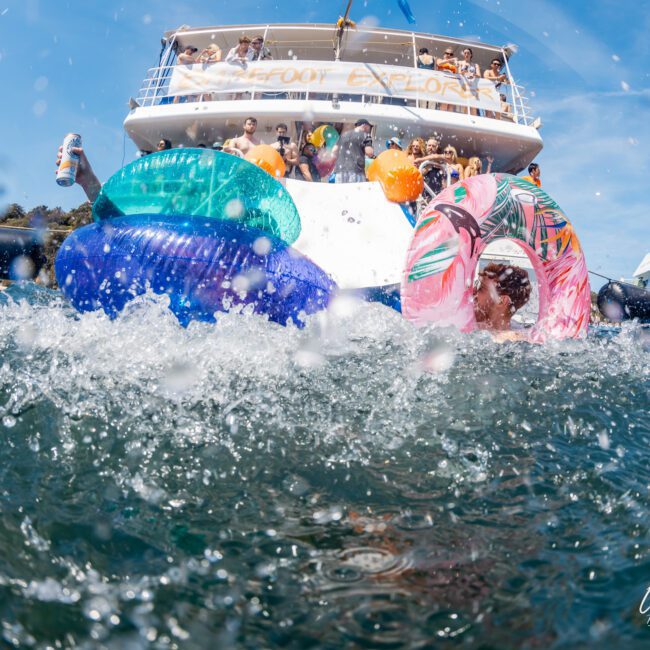 A boat party with people on the deck, pool floats and inflatables in the water, and other boats nearby. Taken from a water-level perspective during an exciting Catamaran party in Sydney.
