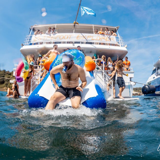 A man in swim trunks slides into the water from a boat slide while others on the luxury yacht enjoy the sunny day. Two large boats are docked side by side, perfect for a Sydney boat party hire.