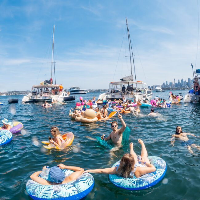 People enjoying a sunny day on the water, floating on inflatable devices near several anchored boats, with a city skyline in the background, perfect for a private yacht charter Sydney Harbour.