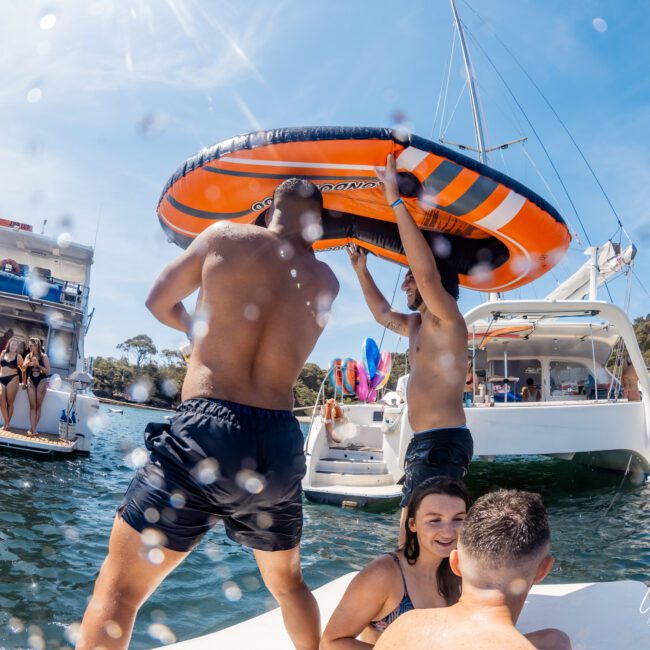 People on boats and in the water on a sunny day. Two men hold an inflatable ring above their heads while others relax nearby, enjoying the vibrant atmosphere of a DJ boat hire Sydney.