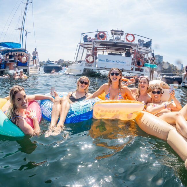 A group of five people relaxing on inflatable floaties in the water, with boats anchored nearby on a sunny day. One boat's sign reads "Rum Runner Cruises." It's the perfect scene for a Sydney boat party hire or a leisurely catamaran party Sydney experience.