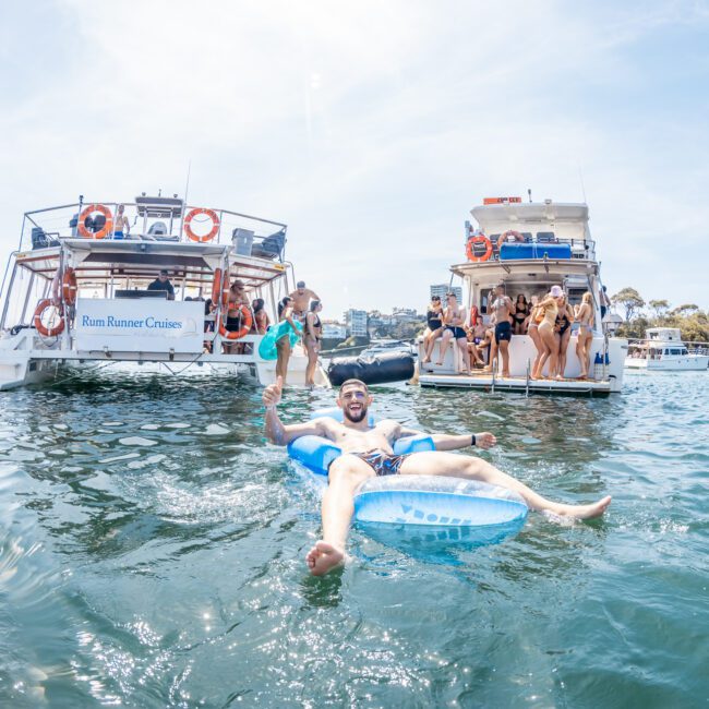 A man relaxes on an inflatable pool float in the water, surrounded by several anchored boats with people socializing aboard, making it the perfect setting for a Sydney boat party hire.