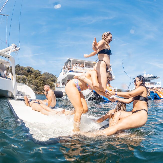 People enjoying water activities between boats on a sunny day, with some sliding into the water and others interacting near the slip and slide at a vibrant Sydney boat party hire.