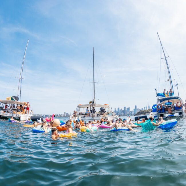 A group of people are floating on inflatables and swimming near moored boats in a large body of water, with a city skyline visible in the background, enjoying what appears to be a luxury yacht hire Sydney event.