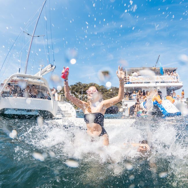 People swimming and splashing water near a boat gathering under a clear sky, with several boats visible in the background, enjoying what could be a luxury yacht hire Sydney experience.