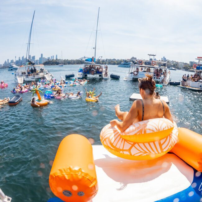 People on inflatable floaties enjoy a sunny day on the water, surrounded by boats and the cityscape. One person is on an orange and white inflatable in the foreground. The idyllic scene sets the perfect backdrop for a Sydney boat party hire or luxury yacht hire Sydney adventure.