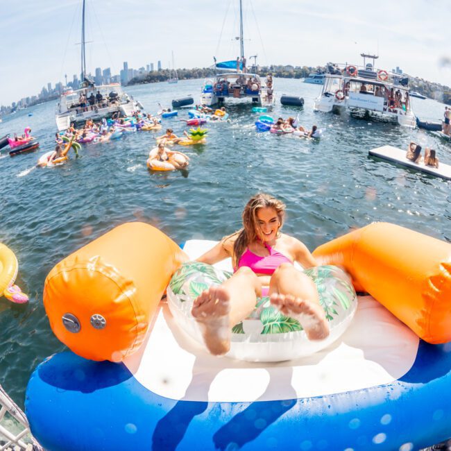 A person relaxes on an inflatable raft in the water as boats and other people on floaties are visible in the background, enjoying a sunny day. Nearby, a private yacht charter Sydney Harbour adds to the scene's allure.