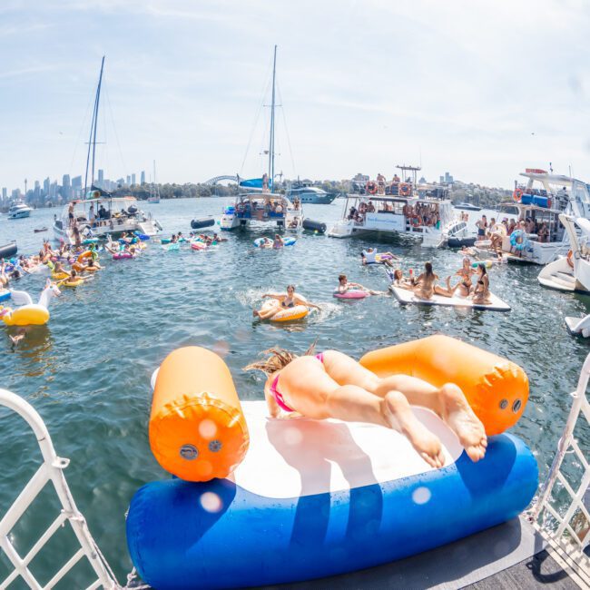 People enjoy a sunny day on a lake with various inflatable floats and boats nearby. Some are swimming, while others relax on boats in the background, including a luxury yacht hire Sydney. Tall buildings can be seen from a distance.