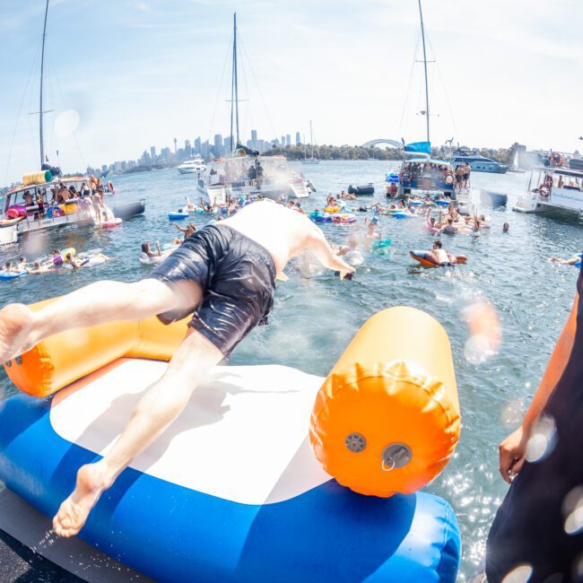 A person wearing a white t-shirt and black shorts jumps from an inflatable platform into a water body with several boats and people floating nearby; a security guard stands on the right. It looks like they are enjoying the festivities of a DJ boat hire Sydney event.
