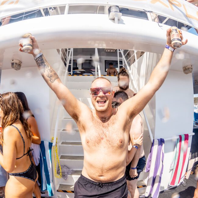 A man with sunglasses raises his arms holding a can in each hand. Several people in swimwear are on a luxury yacht hire Sydney, socializing and holding drinks.