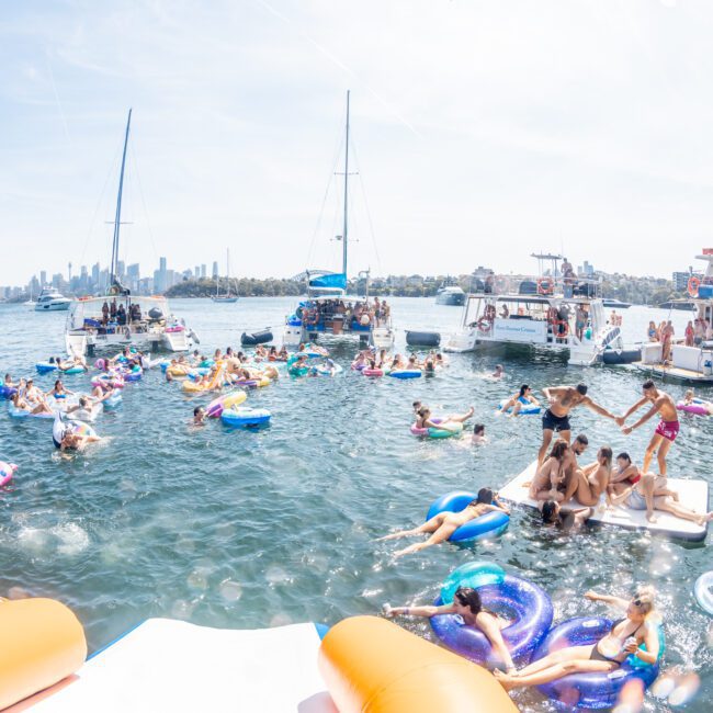 Several people on inflatable floaties enjoy swimming between anchored boats near a city shoreline on a sunny day, possibly part of a lively DJ boat hire Sydney event.