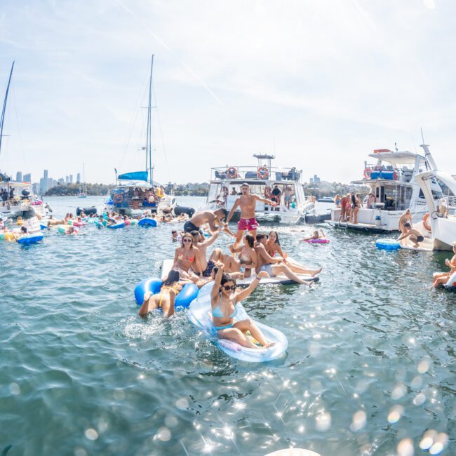 A group of people on inflatable floats enjoy a sunny day on the water, surrounded by several boats. The skyline of a city is visible in the background, making it a perfect setting for corporate boat events Sydney.