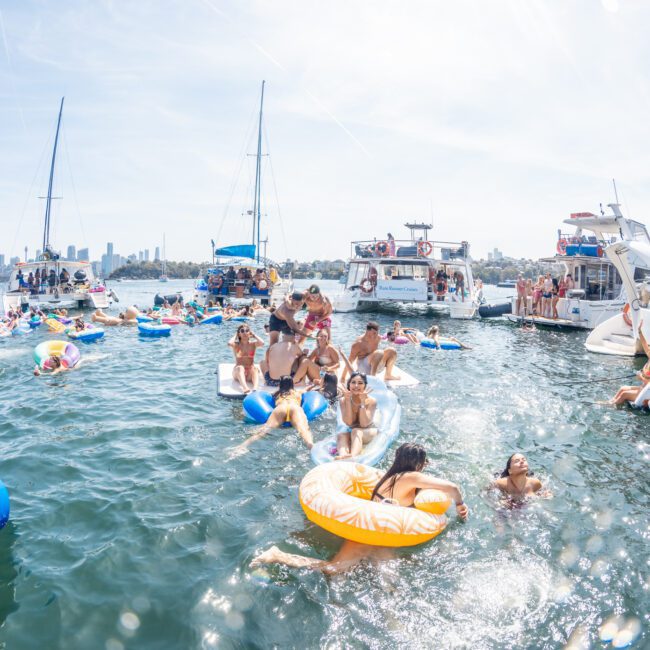 People are enjoying a day on a lake with numerous inflatables and boats. There is a bustling, festive atmosphere as they swim and relax in the water and on the boats, reminiscent of a Sydney boat party hire.