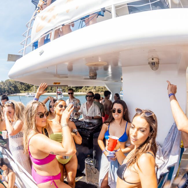 A group of people in swimwear are standing and chatting on the deck of a boat during a lively Sydney boat party hire. Some are holding drinks. Another boat can be seen in the background on the water, enhancing the vibrant atmosphere.