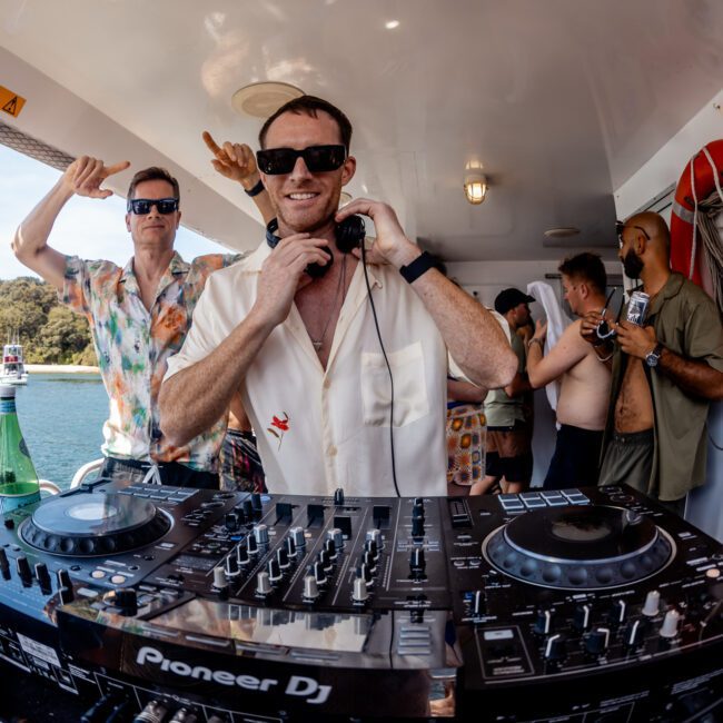 A DJ wearing sunglasses stands behind a Pioneer DJ mixer, surrounded by people celebrating on a boat. The backdrop features a water scene and island, showcasing the vibrant atmosphere of a private yacht charter in Sydney Harbour.