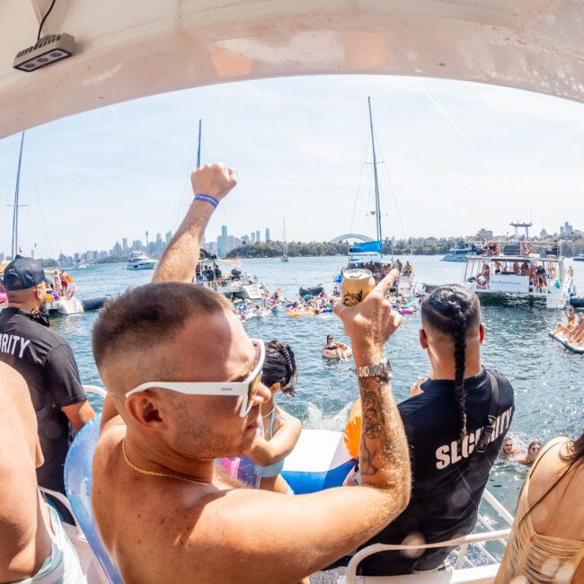 A group of people are celebrating on a boat near a harbor filled with other boats and individuals. One man in sunglasses and a tank top is raising his arm in the foreground, enjoying the festive atmosphere. Consider luxury yacht hire Sydney for your next event for an unforgettable experience.