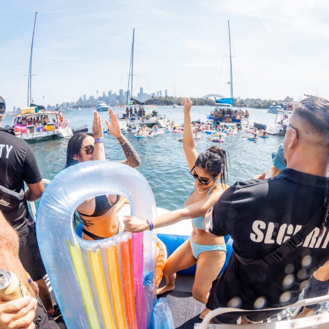 People enjoying a private yacht charter on Sydney Harbour on a sunny day, with some in swimwear, inflatable pool toys, and security personnel present. Boats and the city skyline provide a stunning backdrop.