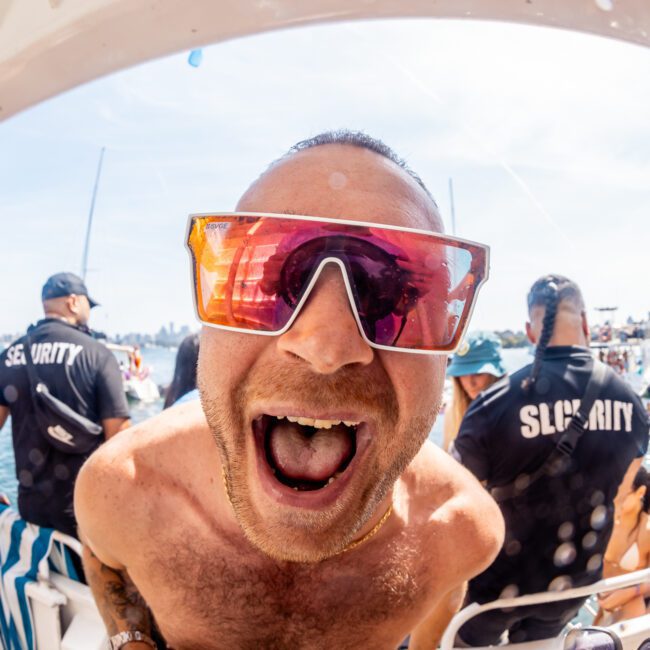 A man in red sunglasses and a gold chain excitedly leans towards the camera on a luxury yacht hire in Sydney, surrounded by people, some wearing matching "Security" shirts.
