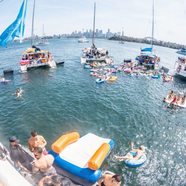 A group of people on boats and inflatables enjoying a sunny day on the water near a cityscape in the background, perfect for a private yacht charter Sydney Harbour or a catamaran party Sydney.