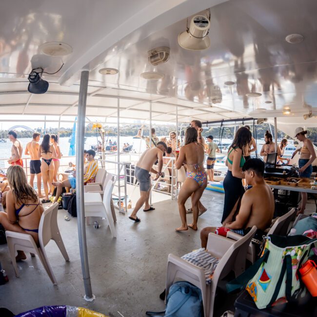 A group of people in swimwear are socializing on a two-level boat under a canopy, with cityscape and water in the background. Some are sitting, others are standing, and various items are scattered around. It captures the essence of a Sydney boat party hire on a private yacht charter in Sydney Harbour.