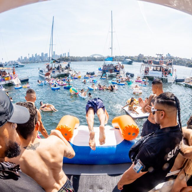 People on a boat gather around as someone dives into the water during a vibrant catamaran party in Sydney, with many other boats and people in floating devices visible on the water.