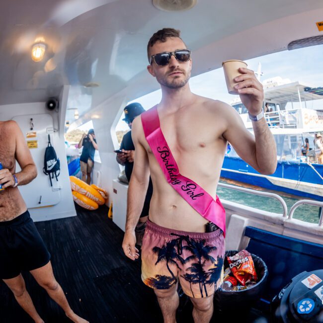 A man wearing sunglasses and a pink sash labeled "Birthday Boy" holds a drink on a catamaran. Another man in swim trunks stands nearby laughing. There's a lively background with people on another boat, enjoying the vibrant atmosphere of Sydney Harbour.