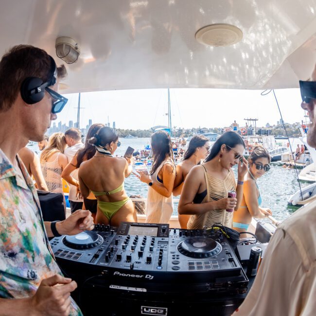 Two DJs are performing on a luxury yacht while a group of people in swimwear enjoys a party on the water, with other boats and the city skyline in the background.