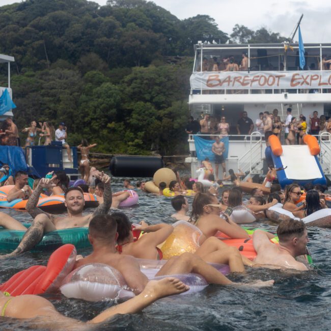 A large group of people floats on inflatable tubes and pool floats in the water near two docked party boats. The boats are crowded with more revelers, and a banner on the larger boat reads "BAREFOOT EXPLORER." Trees and lush greenery create a picturesque backdrop.