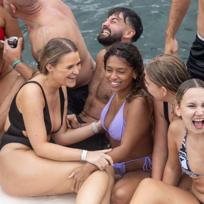 A group of people is sitting close together at the edge of a body of water, wearing swimwear and smiling. They appear to be enjoying a fun and relaxed moment. In the foreground, a young woman in a striped bikini laughs towards the camera, soaking up the beach vibes.