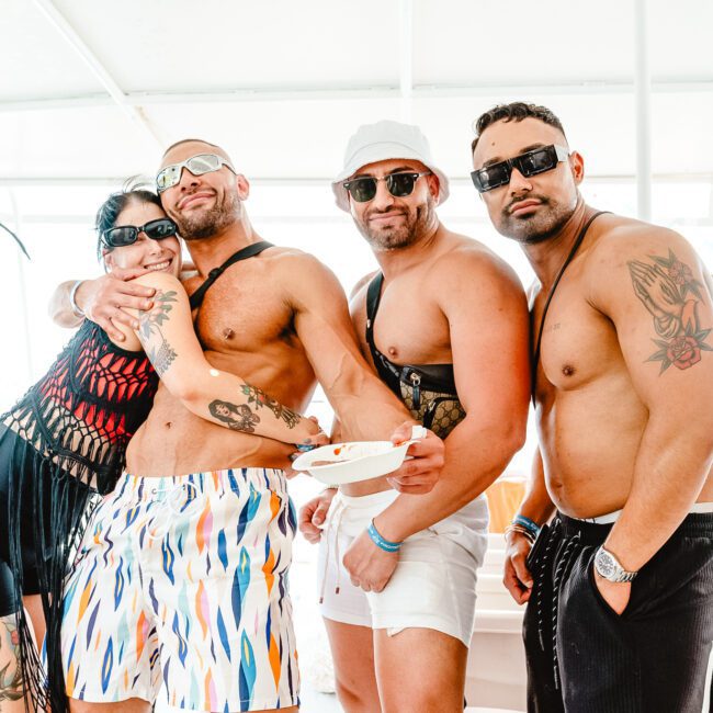 Four friends pose together on a boat in their summer attire. The group includes three men, shirtless in sunglasses and shorts, and one woman happily hugging one of them. The mood is light-hearted and relaxed as they enjoy their sunlit adventure.