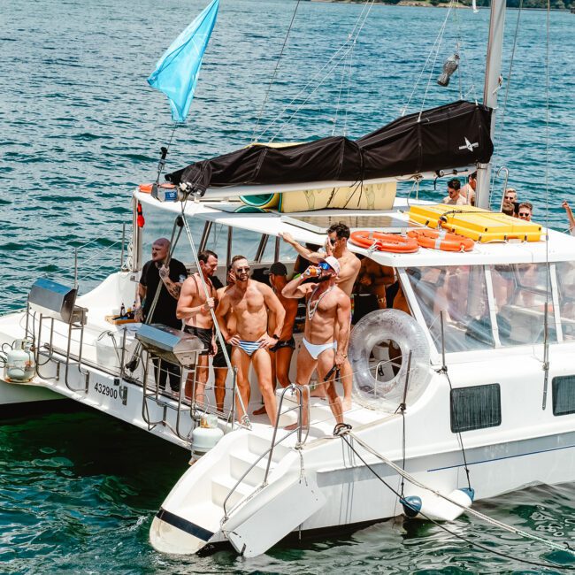 A group of people in swimsuits are gathered on a white catamaran in a sunny, blue ocean. Some stand on the deck, while others sit or lean on the railing. A light blue flag flies at the boat’s stern, and green hills are visible in the background as seagulls fly overhead.
