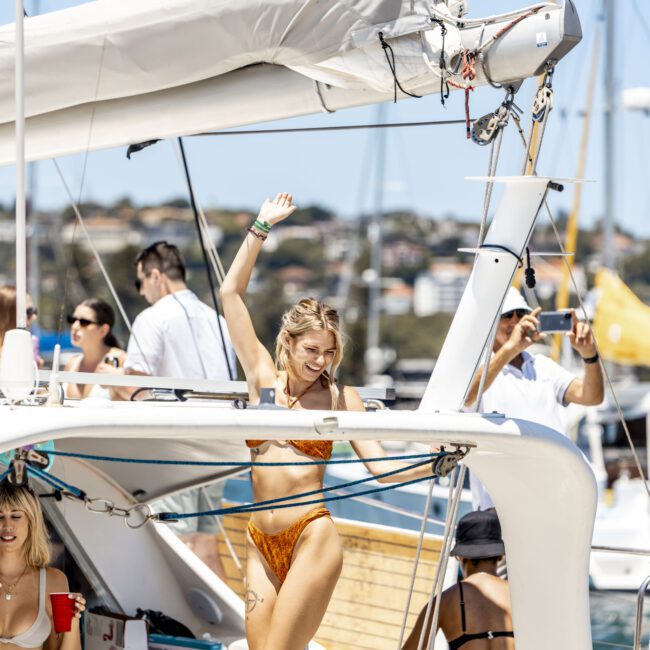 A group of people enjoy a sunny day on a sailboat. In the foreground, a woman in a colorful bikini dances with her arm raised. Other individuals around her engage in conversations and leisure activities, creating a relaxed, cheerful atmosphere against the backdrop of sparkling waters.