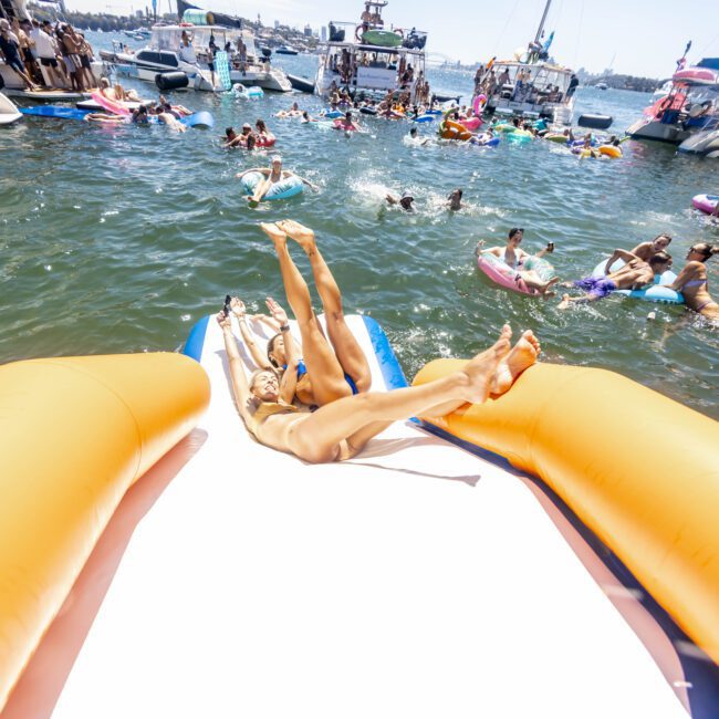 People are sliding down an inflatable slide into the water during a lively boat party. Various boats and people on floaties surround the area, creating a festive and energetic atmosphere under the bright blue sky.