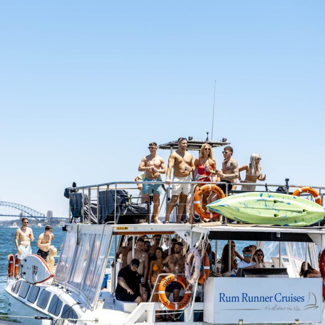 A double-deck boat labeled "Rum Runner Cruises" is crowded with people enjoying a sunny day on the water. Some are on the upper deck, while others paddle kayaks from the lower level. The boat is outfitted with life preservers. A picturesque bridge is visible in the background.