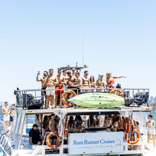 A group of people on the upper deck of "Rum Runner Cruises" boat enjoying a sunny day on the water. Some are posing for a photo while others look out at the scenic skyline. The boat is equipped with a kayak and life rings, adding to the adventurous vibe of the trip.