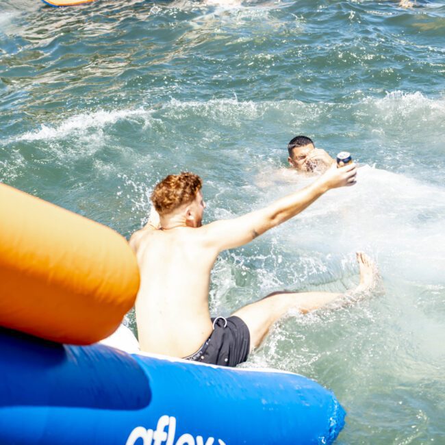 A group of friends are enjoying a sunny day in the crystal-clear blue-green waters. One person is sliding off a vibrant inflatable raft, arm outstretched holding a drink, while others float and swim nearby. The scene is lively and relaxed, capturing the essence of summer fun.