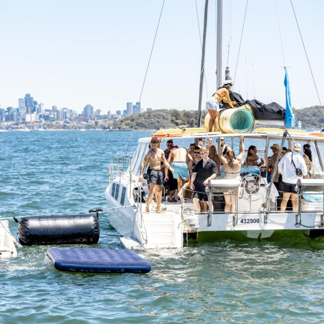 A group of people are gathered on the deck of a catamaran anchored in a bay. Some are standing and others sitting, enjoying the sunny day. A man is swimming near the boat, with an inflatable float nearby. The city skyline and vibrant beach trees create a picturesque backdrop.